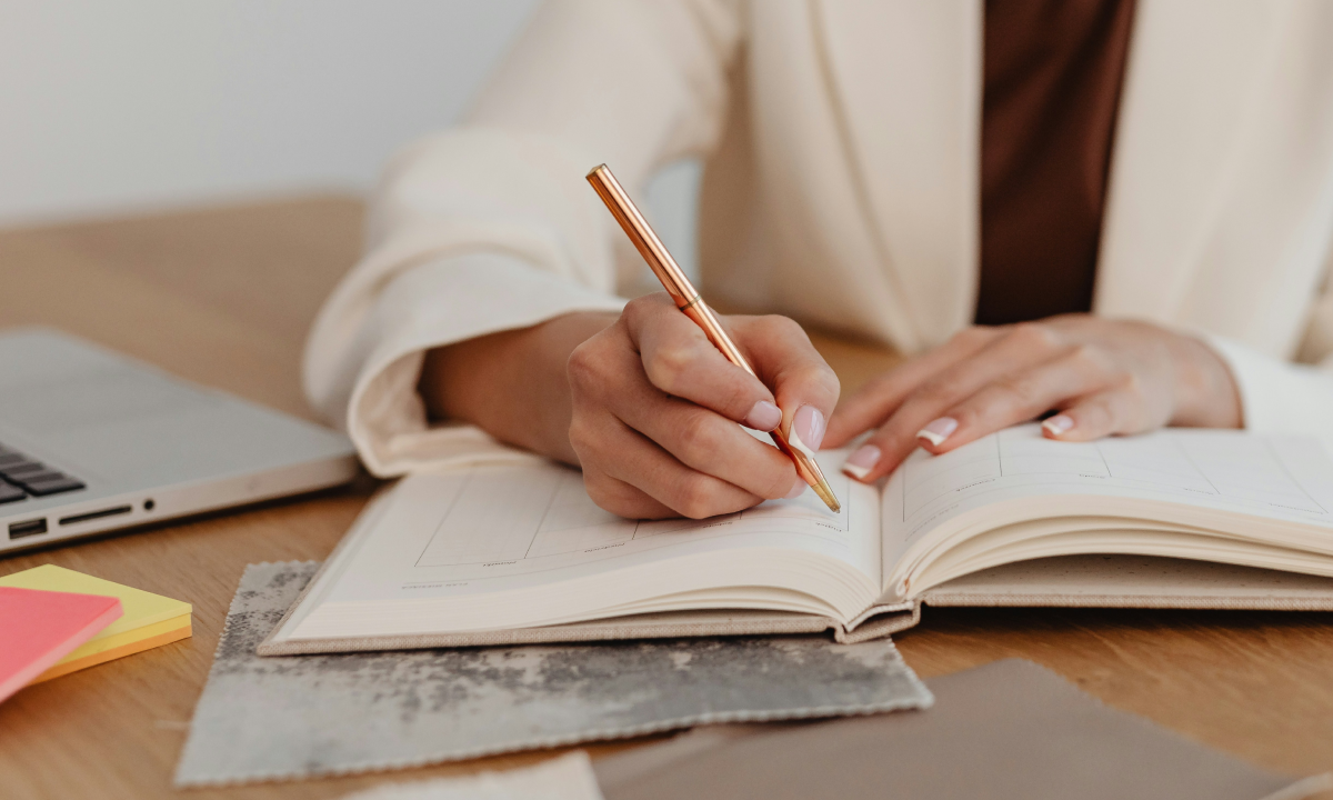 Close-up of a professional writing in a planner at a desk with a laptop and sticky notes, representing strategic performance tracking and marketing planning