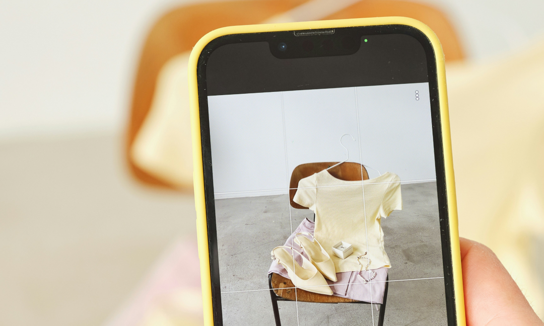 A hand holding a yellow-cased smartphone photographing a styled eCommerce product flat lay of clothing and accessories on a chair, representing paid social media advertising