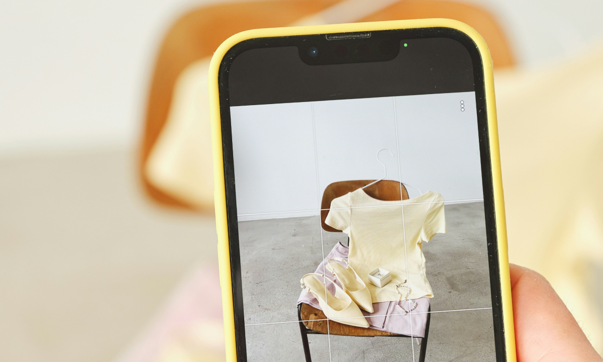 A hand holding a yellow-cased smartphone photographing a styled eCommerce product flat lay of clothing and accessories on a chair, representing paid social media advertising