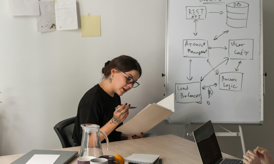 A woman reviewing notes at a conference table in front of a whiteboard with a system architecture diagram, representing strategic planning for scaling businesses